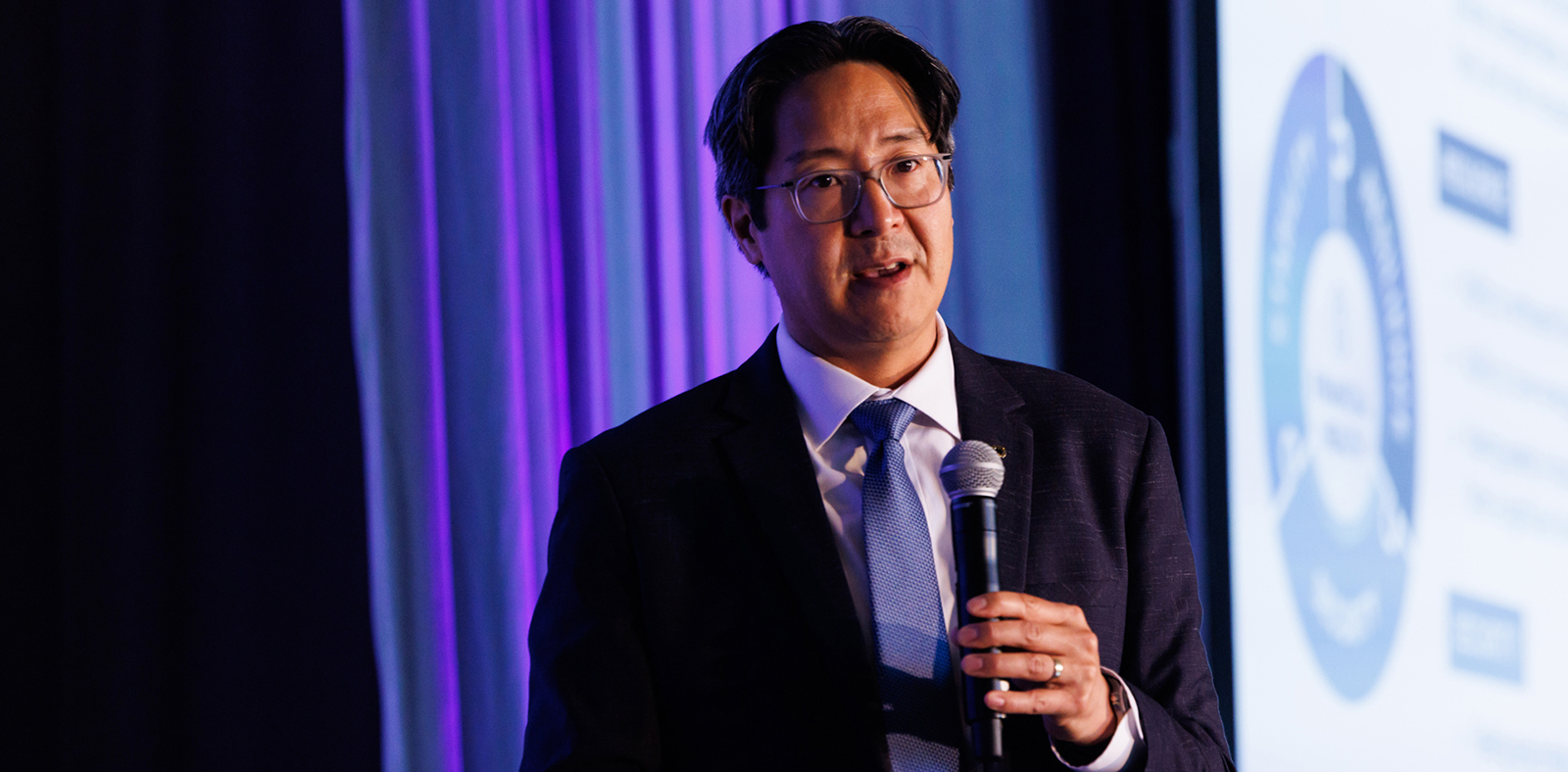 Acting Comptroller Hsu, wearing a dark suit and light blue tie, speaks into a microphone on stage, with purple and blue lighting behind him and a projected slide partially visible in the background. Acting Comptroller Hsu, wearing a dark suit and light blue tie, speaks into a microphone on stage, with purple and blue lighting behind him and a projected slide partially visible in the background.