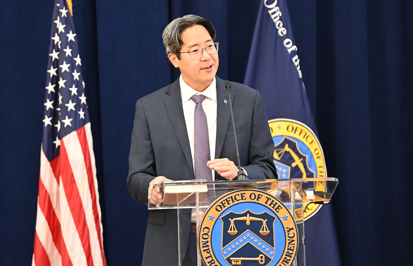 Acting Comptroller Hsu speaks at a clear podium adorned with the Office of the Comptroller of the Currency seal, flanked by the U.S. flag and the OCC flag against a dark blue backdrop. Acting Comptroller Hsu speaks at a clear podium adorned with the Office of the Comptroller of the Currency seal, flanked by the U.S. flag and the OCC flag against a dark blue backdrop.