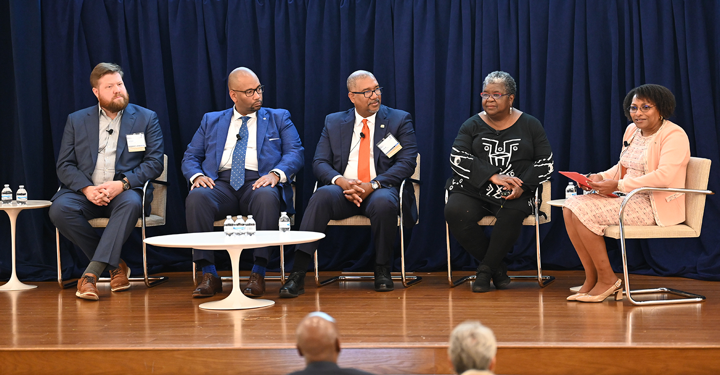 Panelists sit onstage during a discussion at a Project REACh event, with Grovetta Gardineer, the moderator, seated at the far right holding a red folder, against a dark blue backdrop. Panelists sit onstage during a discussion at a Project REACh event, with Grovetta Gardineer, the moderator, seated at the far right holding a red folder, against a dark blue backdrop.
